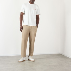 Model wearing Wax London Dean Logo Embroidery T-Shirt in ecru with rust detail, styled with taupe trousers and white trainers.