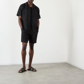 Model wearing Wax London Kurt Linen Shorts in black, styled with matching black linen shirt and dark leather sandals.