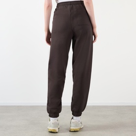 Model wearing Adanola ADA Logo Sweatpants in Coffee Bean, slim-leg with cuffed ankles, shown from behind with white trainers.