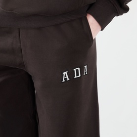 Model wearing Adanola ADA Logo Sweatpants in Coffee Bean, close-up of embroidered ADA logo on slim-leg cotton sweatpants with side pocket.