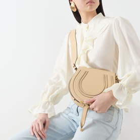 Model carrying Chloe Marcie Saddle Crossbody Bag in Petal Beige, grained leather with gold-toned hardware, worn crossbody with white ruffle blouse