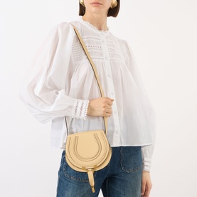 Model carrying Chlo&eacute; Marcie Small Saddle Crossbody Bag in Petal Beige, grained leather, gold-toned hardware, worn crossbody with white broderie