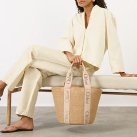 Model carrying Chlo&eacute; Woody Large Basket Tote Bag in Blushy Beige, woven paper finish with logo tape handles, styled with cream wide-leg trousers