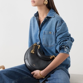 Model carrying Chloe Banana Small Zipped Hobo Bag in black leather with gold-tone hardware, styled with denim shirt and jeans