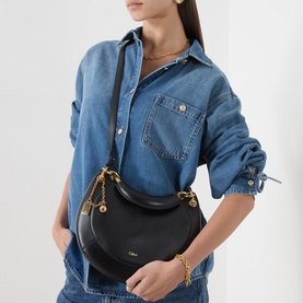 Model carrying Chloe Banana Small Zipped Hobo Bag in black leather, gold-tone hardware and chain detail, worn crossbody over denim shirt