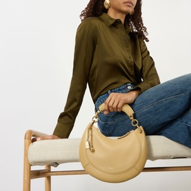 Model carrying Chloe Banana Small Zipped Hobo Bag in Dusty Beige leather, gold-tone hardware, curved silhouette. Styled with olive shirt and jeans.