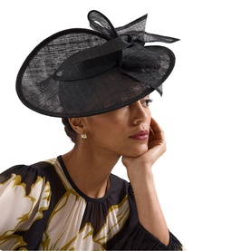 Model wearing Phase Eight black wide-brim sinamay fascinator hat with bow detail, styled with a floral print top and pearl drop earrings.