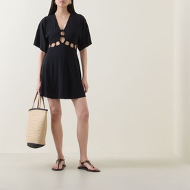 Model wearing Eres Connection Cut-Out Beach Dress in Noir, with V-neck and cut-out detail, styled with a straw tote bag and flat sandals.
