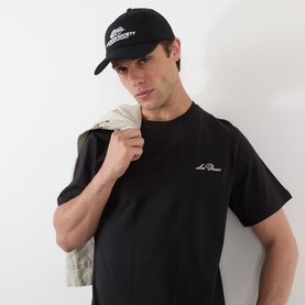 Model with Les Deux Society Dad Cap in black, six-panel cotton cap with embroidered logo, styled with black tee and cream jacket.