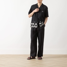Model wearing Les Deux Border Embroidered Shirt in Tap Shoe Black with camp collar and scalloped hem, styled with matching black trousers and sandals.
