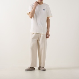 Model wearing Les Deux light ivory cotton T-shirt with small chest logo, styled with cream wide-leg trousers and grey slip-on shoes.