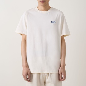 Model wearing Les Deux light ivory cotton T-shirt with small blue chest logo graphic, styled with cream trousers.