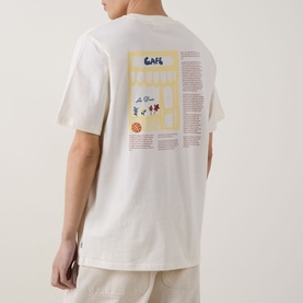Model wearing Les Deux light ivory cotton T-shirt, back view showing yellow and blue graphic print, styled with beige trousers.