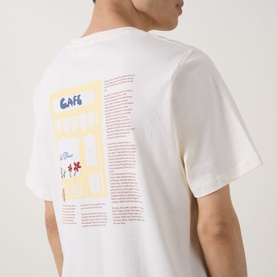 Model wearing Les Deux light ivory cotton T-shirt, back view showing caf&eacute;-themed graphic print with text and floral details.