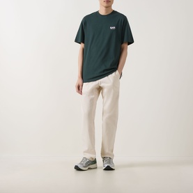 Model wearing Les Deux pine green cotton T-shirt with small chest logo, styled with cream straight-leg trousers and grey trainers.