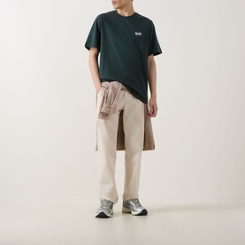 Model wearing Les Deux pine green cotton T-shirt with small chest logo, styled with beige trousers and a tied shirt at the waist.