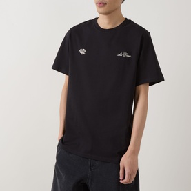Model wearing Les Deux black cotton T-shirt with small chest embroidery details, styled with dark trousers