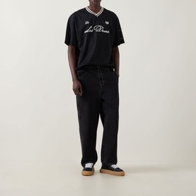 Model wearing Les Deux Logo Mesh Jersey T-Shirt in black with V-neck stripe trim, paired with black trousers and white trainers.