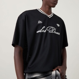 Model wearing Les Deux Logo Mesh Jersey T-Shirt in black with V-neckline, white ribbed trim, script logo embroidery, styled with plaid trousers