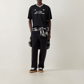 Model wearing Les Deux Logo Mesh Jersey T-Shirt in black with v-neck and logo branding, styled with wide-leg trousers and gum-sole trainers.