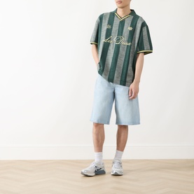 Model wearing Les Deux Stripe Football T-Shirt in pine green with gold stripes, paired with light blue denim shorts and grey trainers.