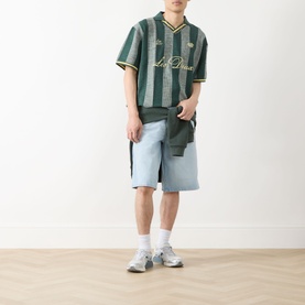 Model wearing Les Deux Stripe Football T-Shirt in pine green with gold logo, styled with light blue denim shorts and grey trainers.