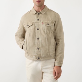 Model wearing Levi's The Trucker Denim Jacket in yellow sand, classic collar, button front, chest flap pockets, red logo tab, with white trousers