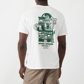 Model wearing Levi's Crew Neck T in Egret, back view showing green "Official Takeover" graphic print, styled with beige trousers.