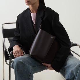 Model carrying Loewe Bilbao Medium Bucket Bag in Dark Chestnut, gold-tone hardware, worn crossbody over black blazer and blue jeans.