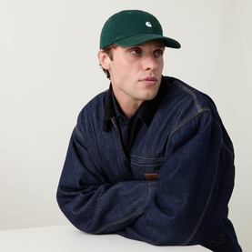Model with Carhartt Madison Logo Baseball Cap in Dark Scarab green, curved visor, white embroidered logo, styled with dark denim jacket