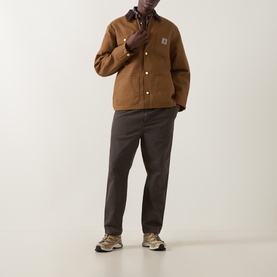 Model wearing Carhartt Hamilton Brown cotton casual jacket with Carhartt patch logo, styled with dark grey trousers and tan trainers.