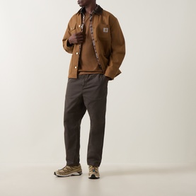 Model wearing Carhartt Casual Trouser in Shungite grey cotton, styled with a tan chore jacket, brown knit and trail trainers.