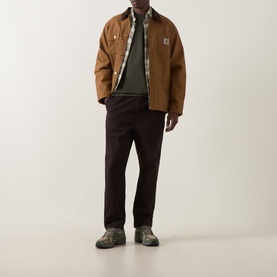 Model wearing Carhartt Casual Trouser in Tobacco brown, styled with a brown canvas jacket and grey-green top, grey trail shoes.