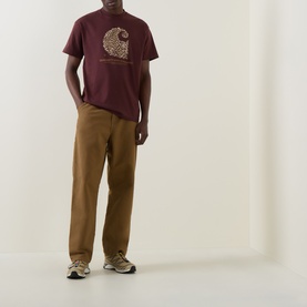 Model wearing Carhartt Entrance Logo Graphic T-Shirt in Amarone, styled with tan wide-leg trousers and grey trainers.