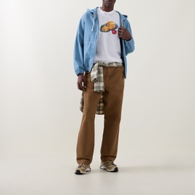 Model wearing Carhartt white crew neck T-shirt with graphic print, styled with light blue zip jacket, plaid shirt and brown trousers.