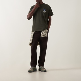 Model wearing Carhartt Crew Neck T in Cypress green with small chest graphic, styled with dark trousers and checked shirt tied at waist.