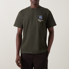 Model wearing Carhartt Crew Neck T in Cypress green with small chest graphic print, paired with dark trousers.