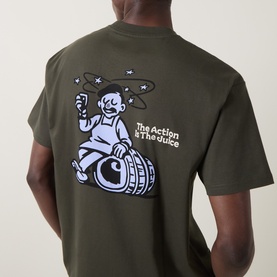 Model wearing Carhartt Crew Neck T in Cypress green, back view showing "The Action & The Juice" graphic print with barrel and character design.