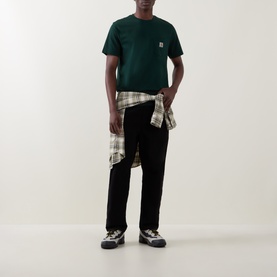 Model wearing Carhartt Crew Neck T in Dark Scarab green with chest pocket, styled with plaid shirt tied at waist and black trousers.
