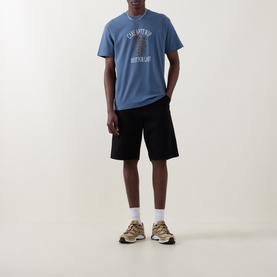 Model wearing Carhartt Crew Neck T in Icesheet blue with chest graphic print, styled with black shorts and tan trainers.
