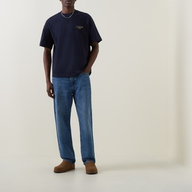 Model wearing Carhartt Signature Script Logo T-Shirt in Dark Navy, styled with mid-blue straight-leg jeans and tan suede shoes.