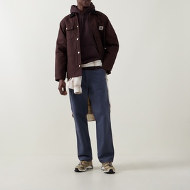 Model wearing Carhartt hoodie in Palisander/Gold layered under brown jacket, styled with navy trousers and trail trainers