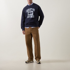 Model wearing Carhartt Dark Navy cotton sweatshirt with collegiate graphic print, styled with tan trousers and white shirt underneath.
