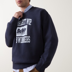 Model wearing Carhartt dark navy cotton sweatshirt with large Big WIP Winners graphic print, layered over a light blue shirt.