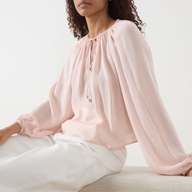 Model wearing Frame Denim Taffy silk blouse with gathered neckline, tie detail and pleated long sleeves, styled with white trousers