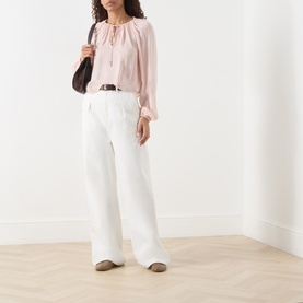 Model wearing FRAME DENIM Taffy silk blouse with white wide-leg trousers, brown belt, and carrying a dark shoulder bag.