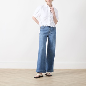 Model wearing FRAME DENIM white short-sleeve shirt with broderie anglaise hem, styled with wide-leg blue jeans and black flat sandals.