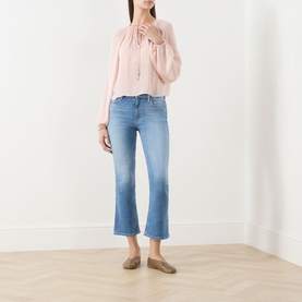 Model wearing Paige Crop Boot jeans in Cinerama blue denim with raw hem, styled with a pink blouse and taupe flat mules.