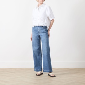 Model wearing Paige Encino straight jeans in mid-blue denim, styled with a white broderie anglaise top and tan sandals.