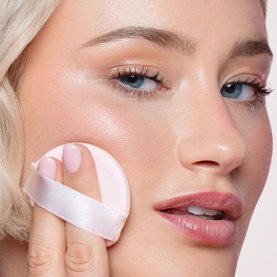 Model wearing Sculpted by Aimee Cream Cushion Foundation Refill, applying compact cushion applicator to cheek for a glowing finish.